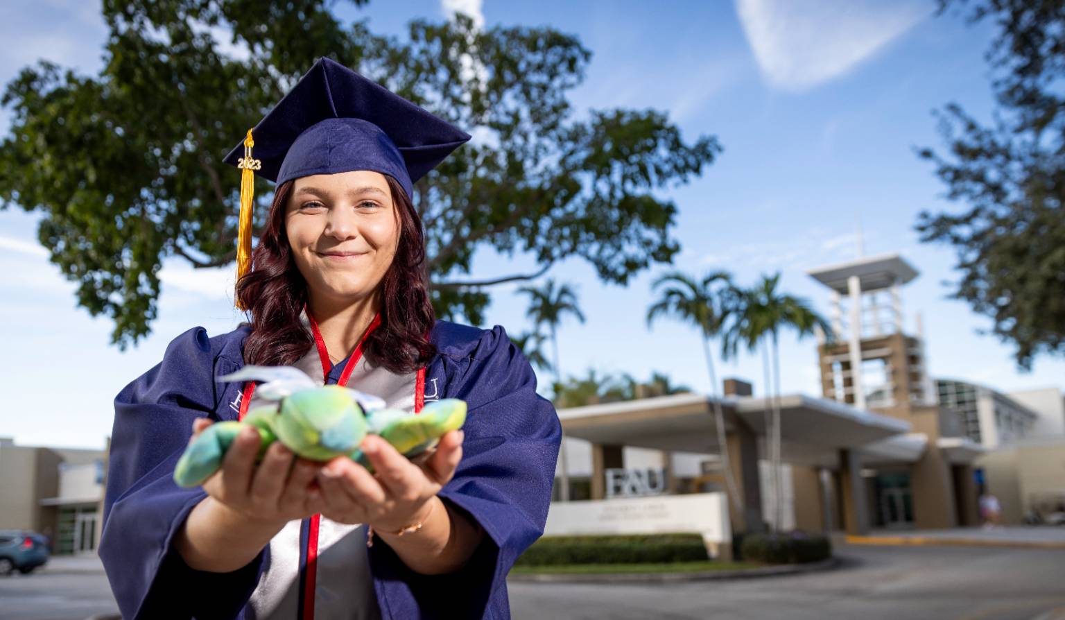 Graduate in cap and gown smiling while holding a plush sea turtle, standing outdoors at Florida Atlantic University with palm trees and modern buildings in the background.
