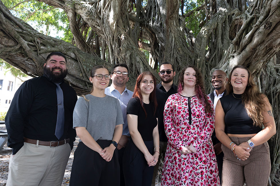 Eight people pose in front of a large tree smiling at the camera.