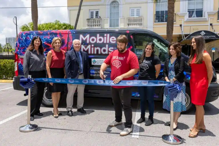 Background includes a blue van with a red brain and the world "Mobile Minds" in white letters. In front of it are 6 adults wearing business attire. At the forefront of the photo is a long blue ribbon with 'Florida Atlantic University' across the ribbon. A man wearing a red shirt and jeans is cutting the ribbon with a pair of scissors.