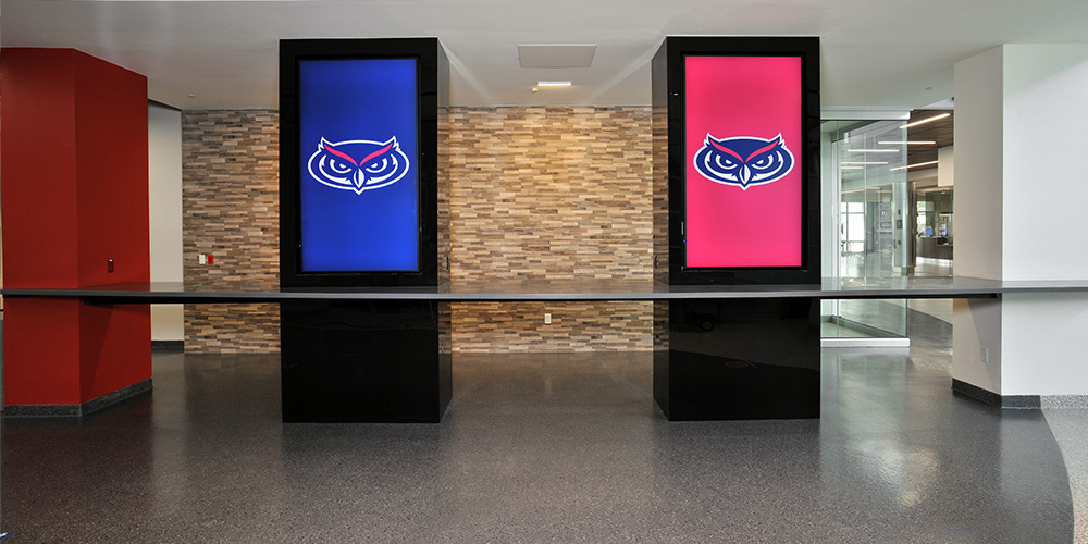 Interior lobby with two tall digital kiosks displaying owl logos on blue and red screens against a stone wall