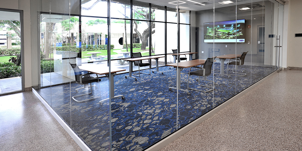 Coconut Palm Room event space at the FAU Student Union, featuring a glass-enclosed room with movable tables and chairs, patterned blue carpet, wall-mounted display screen, and large windows overlooking an outdoor courtyard