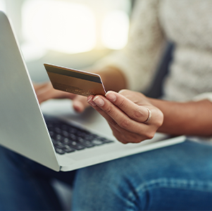 Woman with a laptop on her lap holding up a credit card. Her face is not visible.