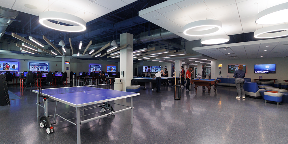 Open esports and gaming center with a ping-pong table, pool tables, rows of gaming computers, and students using the space under modern overhead lighting